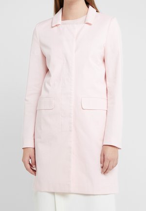 Una donna che indossa un cappotto lungo rosa pallido con tasche a soffietto sul davanti, sopra pantaloni bianchi, in piedi contro uno sfondo chiaro e semplice.