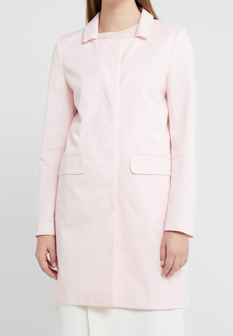 Nainen, joka pukeutuu vaaleanpunaiseen pitkähihaiseen takkiin, jossa on etupussit, valkoisten housujen päällä, seisoo tasaista vaaleaa taustaa vasten.