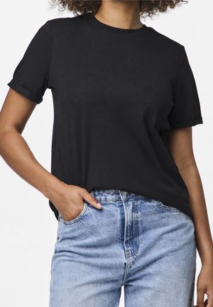 Kobieta w prostym czarnym t-shircie z krótkim rękawem, lekko wsadzonym w jasnobłękitne dżinsy z wysokim stanem, z jedną ręką w kieszeni.