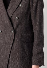 Veste croisée marron foncé à motif chevron avec boutons décoratifs argentés et une poche à rabat à l'avant côté droit.