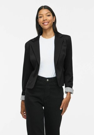 Jeune femme aux longs cheveux foncés portant un blazer noir sur une chemise blanche et un pantalon noir, souriante sur un fond blanc uni.