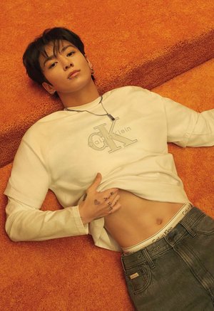 Young man with dark hair and piercings lies on orange carpet, lifting white Calvin Klein shirt to reveal toned abdomen and tattooed hand.
