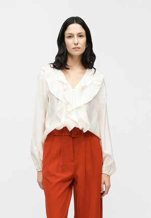 Hvid bluse med flæser foran og lange ærmer, kombineret med højttaljede røde bukser med bælte. Glat, letvægtsstof.