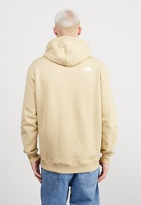Sudadera con capucha beige de textura suave, que presenta un logo blanco en la parte posterior cerca del hombro derecho, combinada con vaqueros azules.