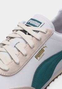 Κοντινό πλάνο άσπρου αθλητικού παπουτσιού Puma Arizona με τιρκουάζ ρίγα, μπεζ σουέντ λεπτομέρειες, λευκά κορδόνια και χρυσό λογότυπο Puma Arizona στο πλάι.