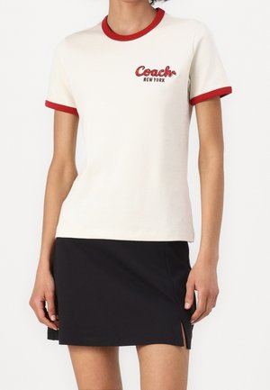 Kremowy T-shirt z krótkim rękawem z czerwonym obszyciem i haftowanym logo "Coach New York". Połączony z czarną spódnicą z bocznym rozcięciem.
