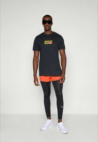 Black t-shirt with "BERLIN RUN" in yellow, orange shorts, black leggings, and white sneakers. Accessories include dark sunglasses.