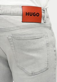 Ανοιχτό γκρι τζιν τσέπη στην πλάτη με ορθογώνιο πορτοκαλί δερμάτινο ταμπελάκι με την ένδειξη "HUGO" ραμμένο πάνω από την τσέπη.