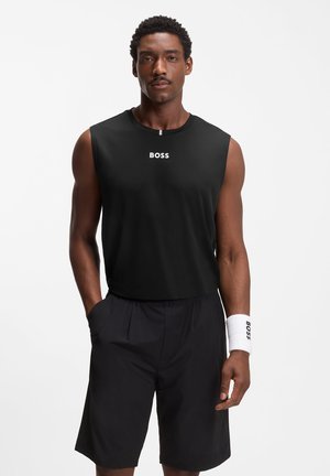 Uomo in piedi che indossa una maglietta nera senza maniche e pantaloncini con fascia bianca al polso, logo "BOSS" sulla maglietta e sulla fascia, mani nelle tasche.