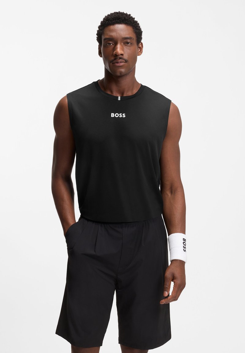 Άνδρας που στέκεται, φορά μαύρο αμάνικο μπλουζάκι και σορτς με λευκό βραχιόλι στον καρπό, λογότυπο "BOSS" στο μπλουζάκι και το βραχιόλι, τα χέρια στις τσέπες.