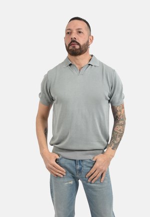 Uomo con barba e piercing all'orecchio indossa una polo grigio chiaro a maniche corte e jeans blu strappati, mostrando un tatuaggio floreale colorato sul braccio sinistro.