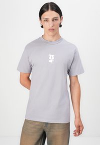 Lys grå bomulds t-shirt med korte ærmer, rund halsudskæring og et hvidt grafisk logo centreret på brystet. Glat tekstur.