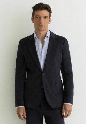 Joven con cabello corto oscuro vestido con una chaqueta de traje oscura sobre una camisa de rayas azul clara, de pie frente a una pared blanca y lisa.