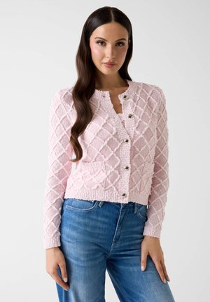 Femme aux longs cheveux foncés portant un cardigan en maille texturée rose avec des boutons argentés et un jean bleu, debout devant un fond uni.
