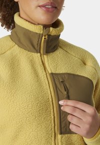 Fleece jack in het geel met een hoge kraag. Voorzien van een ritsborstas in bruin. Textuur, warme stof met een zachte afwerking.