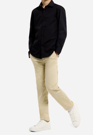 Mand iført en sort skjorte med knapper, beige bukser, hvide sokker og hvide sneakers, stående med hænderne i lommerne, højre ben krydset over venstre.