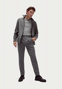 Camicia grigia chiaro con bottoni e colletto, indossata sopra una t-shirt grigia, abbinata a pantaloni grigi su misura e scarpe nere. Tessuto liscio.