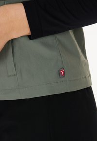 Zelená lehká bunda s vysokým límcem, která má boční zip a malou červenou značku na lemu. Pod ní je viditelná černá látka.