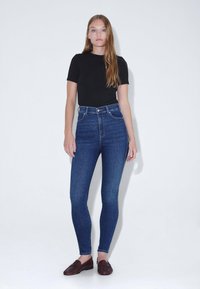 Jeans ajustados de tiro alto en azul con una textura suave, combinados con una camiseta negra ajustada y mocasines marrón oscuro.