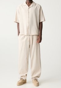 Man in een beige overhemd met korte mouwen, bijpassende losse broek met een trekkoord en beige veterschoenen op een effen witte achtergrond.