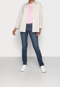Camisa de botones color beige claro, camiseta rosa por debajo, jeans azul oscuro y zapatillas blancas. Presenta un corte relajado y mangas largas.