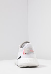 Zapatilla deportiva blanca con parte superior de malla, acentos grises y un contra talón negro. Presenta una entresuela Boost expuesta y una pequeña etiqueta de logo.