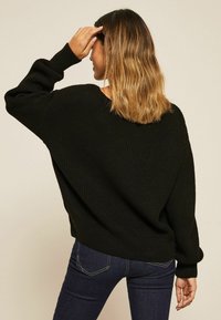 Pull en maille noir avec une coupe décontractée, des épaules tombantes et des poignets côtelés. Associé à un jean en denim foncé. Le tissu a une apparence texturée.