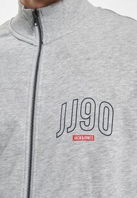 Sivá bundová mikina na zips s vysokým golierom. Výrazné prvky zahŕňajú potlač „JJ90“ tmavomodrou farbou a „JACK & JONES“ červenou farbou na ľavej strane hrudníka.