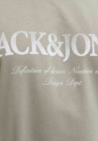 Ανοιχτό μπεζ ύφασμα με λευκό κείμενο "JACK&JONES" σε μεγάλο μέγεθος γραμματοσειράς και μικρότερο καλλιγραφικό κείμενο που γράφει "Definition of denim Nineteen n Design Dept."