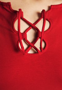 Top rojo con escote redondo que presenta un detalle de cordones cruzados. Tela suave con una forma ajustada. Acentos de textura contrastante.