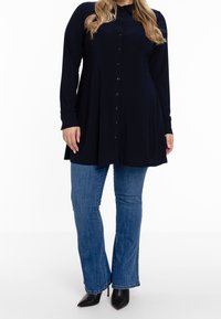 Abito camicia a maniche lunghe di colore navy con bottone frontale, orlo curvo e tessuto morbido, abbinato a jeans a zampa blu e décolleté nere con punta.