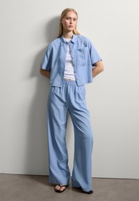 Hellblaues kurzärmeliges Hemd mit einer Tasche, kombiniert mit lässigen hellblauen Hosen mit elastischem Bund. Schwarze Sandalen.