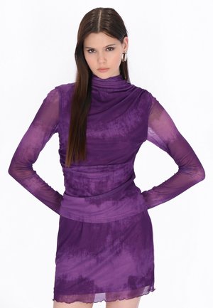 Tânără cu păr lung și castaniu poartă o rochie mov transparentă, mulată, stând cu mâinile în șolduri pe un fundal alb.