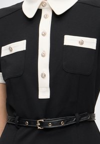 Μαύρο φόρεμα με λευκή γιακά, λευκό κρίκο κουμπιών, πτερύγια τσέπης, κουμπιά με πολύτιμους λίθους και μαύρη ζώνη με χρυσή αγκράφα στη μέση.