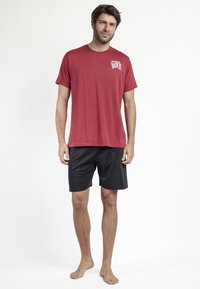 Camiseta roja de manga corta con un logo en el pecho, combinada con pantalones cortos negros. Ambas prendas están hechas de una tela suave y transpirable.
