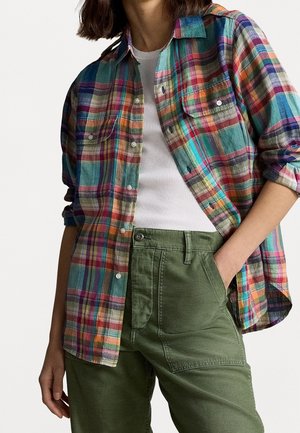 Frau trägt ein mehrfarbig kariertes Hemd über einem weißen Tanktop und olivgrüne Hosen, eine Hand in der Tasche, vor einfachem Hintergrund.