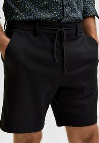 Černé šortky vyrobené z hladkého, měkkého materiálu. Mají elastický pas se šňůrkou a boční kapsy. Jednoduchý, přizpůsobený design bez vzorů.