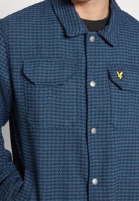 Giacca a camicia a quadri blu con due tasche sul petto, bottoni a pressione in metallo e un piccolo logo giallo sulla tasca sinistra. Tessuto morbido.