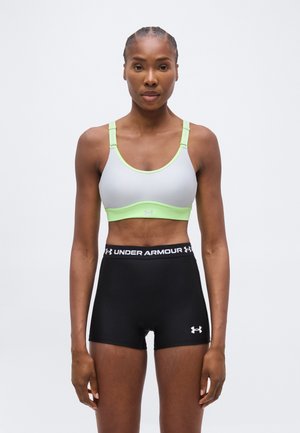 Kvinde stående front mod kamera iført en hvid og grøn sports-bh og sorte Under Armour træningsshorts mod en ensfarvet hvid baggrund.