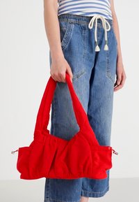 Sac à main en tissu rouge avec une texture douce, design plissé, poignées doubles et une poche zippée, tenu par une personne portant un jean bleu.