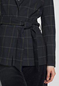 Granatowa marynarka w kratę z żółtymi i białymi detalami przeszycia, z paskiem w talii. Charakteryzuje się fakturowaną tkaniną, wyciętym kołnierzem i klapą.