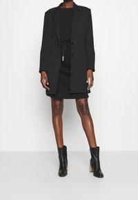 Blazer negro con dos botones, usado sobre una blusa negra y una falda ajustada. Botines negros de tacón alto. Textura de tela suave.