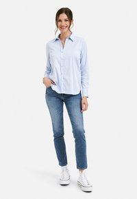 Light blue button-up shirt with long sleeves, paired with blue jeans and white sneakers; relaxed fit with a rounded hem.