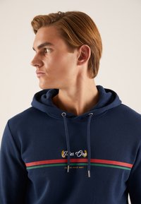 Temno modra kapuca z logotipom, vodoravnimi rdečimi in zelenimi črtami ter srebrnimi dodatki na vrvicah.