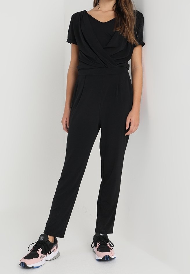 Femme portant un haut cache-cœur noir à manches courtes, un pantalon noir taille haute et des baskets noires et roses, debout devant un fond uni.