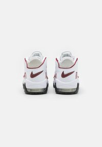 Vita sportskor i vitt med röda accenter, en texturerad häl, synlig luftdämpning och en gummisula; har en framträdande logotyp på baksidan.