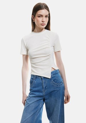 Witte geribbelde top met korte mouwen en plooien aan de zijkant, met een asymmetrische snit. Gecombineerd met hoge blauwe denim jeans.