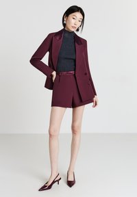 Donna che indossa un blazer bordeaux e pantaloni corti abbinati, con un maglione nero a collo alto e décolleté bordeaux.
