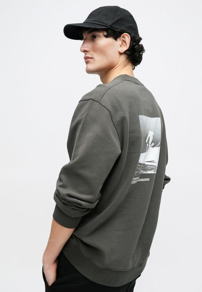 Φούτερ σε ελιά πράσινο με στρογγυλή λαιμόκοψη και ριμπ μανσέτες, με γραφικό σχέδιο στην πλάτη. Μαλακή υφή, άνετη γραμμή. Φοράει μαύρο καπέλο.