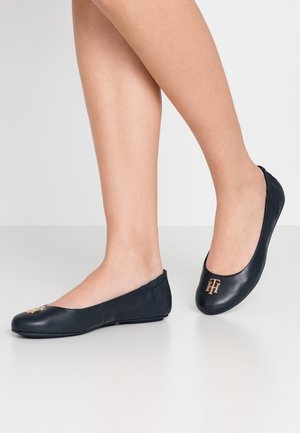 Voeten met zwarte leren ballerina's met gouden "TH"-logo's op de tenen, geposeerd tegen een effen witte achtergrond.
